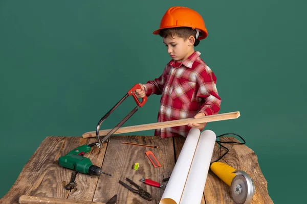 Retrato de menino bonito, criança na imagem do construtor, designer em capacete de proteção laranja usando ferramentas de trabalho isolado no fundo verde — Fotografia de Stock