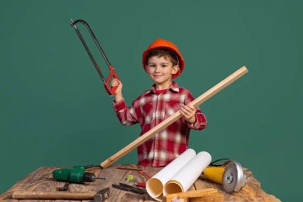 Retrato de niño lindo, niño en la imagen del constructor, diseñador en casco protector naranja utilizando herramientas de trabajo aisladas sobre fondo verde — Foto de Stock