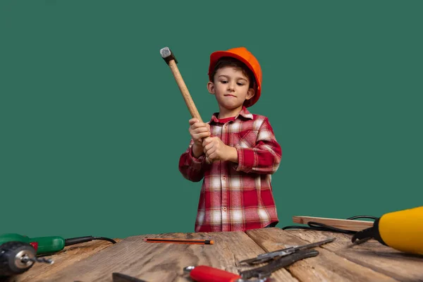 Retrato de niño lindo, niño en la imagen del constructor, diseñador en casco protector naranja utilizando herramientas de trabajo aisladas sobre fondo verde — Foto de Stock