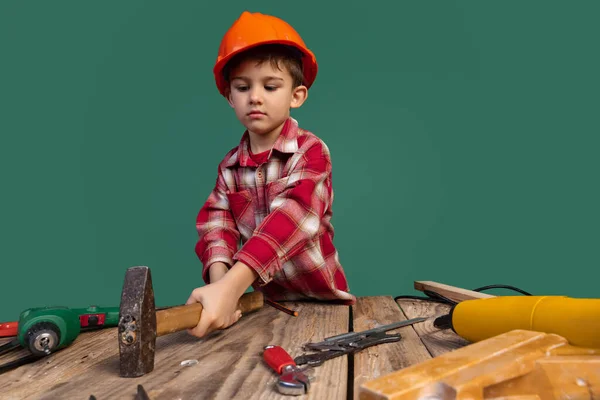 Retrato de niño lindo, niño en la imagen del constructor, diseñador en casco protector naranja utilizando herramientas de trabajo aisladas sobre fondo verde — Foto de Stock