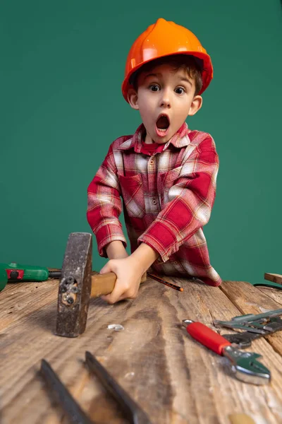 Retrato de niño lindo, niño en la imagen del constructor, diseñador en casco protector naranja utilizando herramientas de trabajo aisladas sobre fondo verde — Foto de Stock