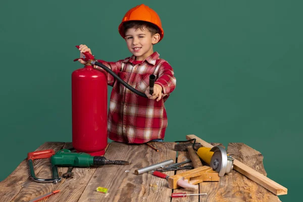 Şirin küçük bir çocuğun portresi, inşaatçı görüntüsü, turuncu koruyucu kasklı tasarımcı yeşil arka planda izole edilmiş iş aletleri kullanıyor. — Stok fotoğraf