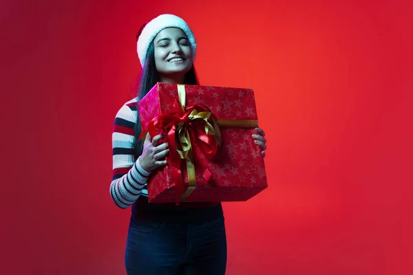 Retrato de la joven chica feliz en ropa de abrigo y sombrero de Navidad celebración de caja de regalo enorme aislado en fondo de estudio de color rojo — Foto de Stock