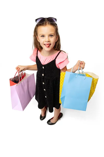 Студійний знімок досить маленької милої дитини, що стоїть з різнокольоровими пакетами покупок ізольовані на білому тлі . — стокове фото