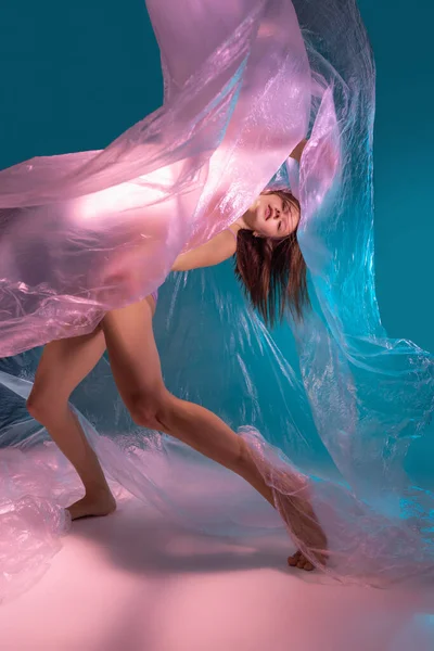 Динамический портрет молодого гибкого танцора, танцующего с тканью, выделенной на градиентно-голубом фоне в неоновом свете — стоковое фото
