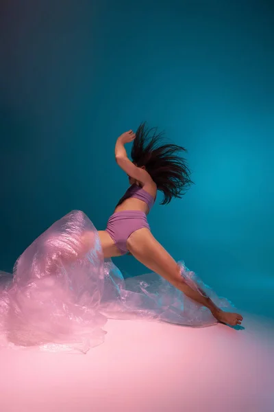 Одна молодая гибкая танцовщица танцует с тканью, выделенной на градиентном синем фоне в неоновом свете — стоковое фото