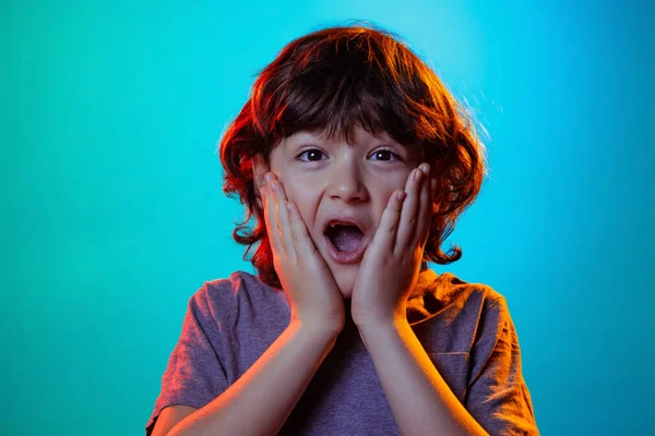 Muotokuva pieni poika, kihara yllättynyt lapsi seisoo suu auki eristetty sininen studio tausta neon valo, suodatin. — kuvapankkivalokuva