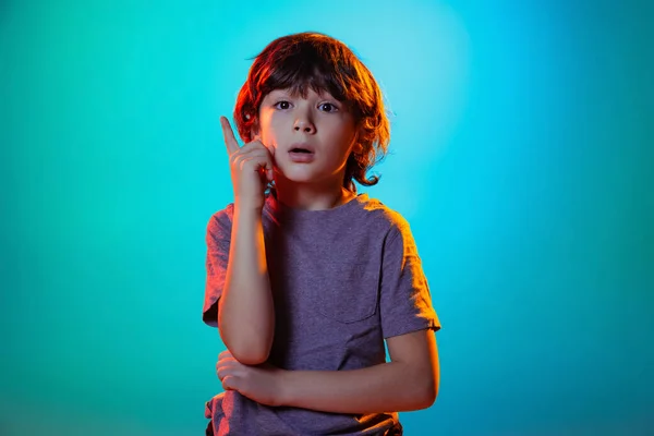 Μισού μήκους πορτραίτο του μικρού αγοριού, παιδί που σηκώνει το δάχτυλο επάνω απομονωμένο σε μπλε φόντο στούντιο σε νέον φως, φίλτρο. — Φωτογραφία Αρχείου