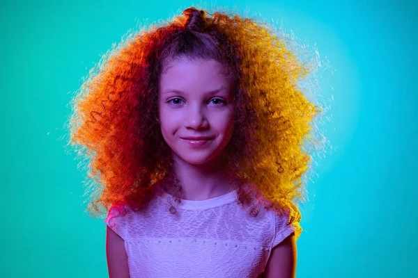 Close-up kleine schattig krullend kleuterschool mooi meisje kijken naar camera geïsoleerd op blauwe studio achtergrond in paars neon licht. — Stockfoto