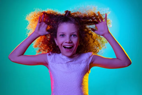 Close-up kleine schattig krullend kleuterschool mooi meisje kijken naar camera geïsoleerd op blauwe studio achtergrond in paars neon licht. — Stockfoto