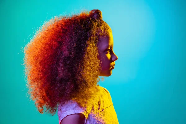 Vista del perfil de la pequeña niña hermosa preescolar rizada posando aislada sobre fondo de estudio azul en luz de neón púrpura. — Foto de Stock