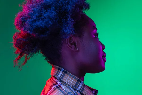 Vista laterale della giovane ragazza dalla pelle scura con adorabile acconciatura isolata su sfondo verde scuro studio in luce viola neon. — Foto Stock