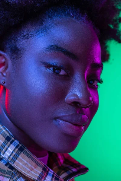 Nahaufnahme des Gesichts eines dunkelhäutigen jungen Mädchens, das isoliert auf dunkelgrünem Studiohintergrund in lila Neonlicht in die Kamera blickt. — Stockfoto