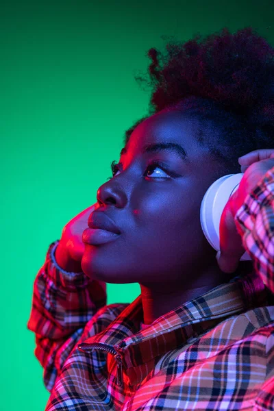 Close-up szczęśliwa młoda dziewczyna, student słuchający muzyki w słuchawkach odizolowanych na ciemnozielonym tle studio w fioletowym świetle neonu. — Zdjęcie stockowe