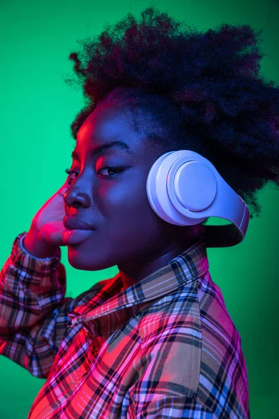 Muchacha joven de cerca, estudiante escuchando música en auriculares aislados sobre fondo de estudio verde oscuro en luz de neón púrpura. —  Fotos de Stock