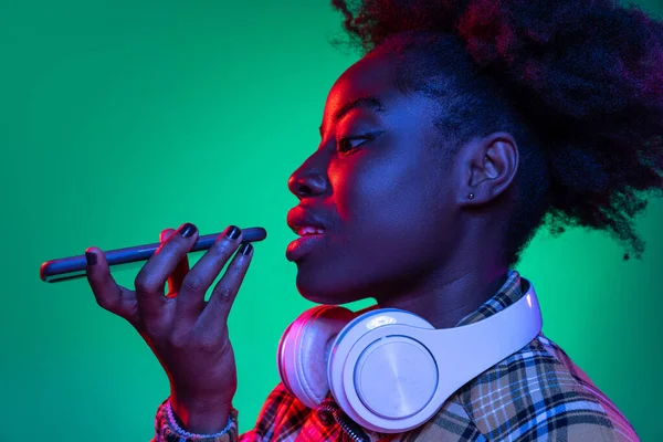 Close-up giovane bella ragazza africana, studente utilizzando il telefono isolato su sfondo verde scuro studio in luce viola al neon. — Foto Stock