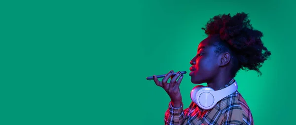 Broşür genç, güzel Afrikalı kız, telefon kullanan öğrenci koyu yeşil stüdyo arka planında, mor neon ışıkla izole edilmiş.. — Stok fotoğraf