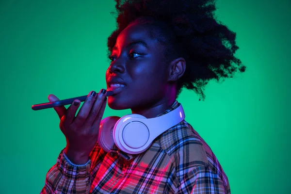 스튜디오는 보라색 네온 조명을 받은 어두운 녹색 스튜디오 배경에서 고립된 핸드폰을 사용하는 젊은 아프리카 소녀의 사진을 찍었습니다.. — 스톡 사진