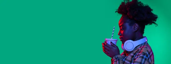 Flyer junge hübsche afrikanische Mädchen, Student Limonade trinken isoliert auf dunkelgrünem Studiohintergrund in lila Neonlicht. — Stockfoto
