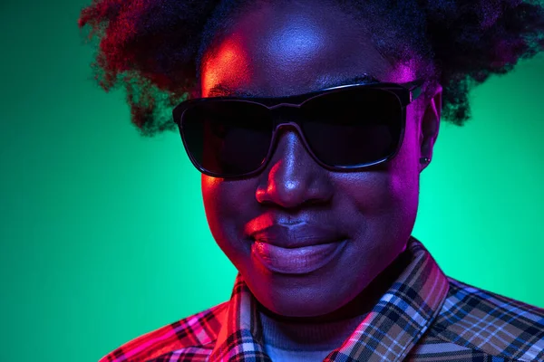 Nahaufnahme des Gesichts eines dunkelhäutigen jungen Mädchens mit Sonnenbrille isoliert auf dunkelgrünem Studiohintergrund in lila Neonlicht. — Stockfoto