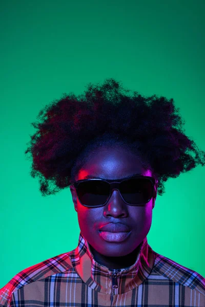 Portrait of sad African young girl in sunglasses posing isolated on dark green studio background in purple neon light. — Stok Foto