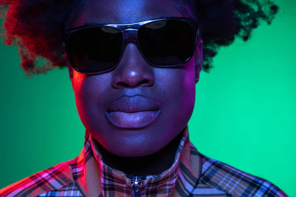 Nahaufnahme des Gesichts eines dunkelhäutigen jungen Mädchens mit Sonnenbrille isoliert auf dunkelgrünem Studiohintergrund in lila Neonlicht. — Stockfoto