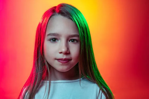 Close-up kleuter meisje, rustig kind met lang glanzend haar op zoek naar camera geïsoleerd op rood gele studio achtergrond in neon licht, filter. — Stockfoto