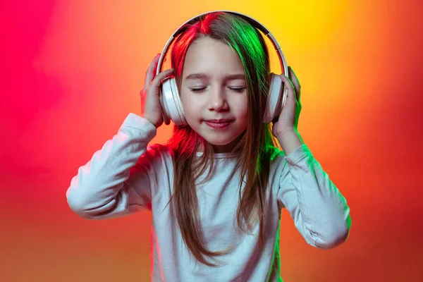 Портрет дошкольницы, счастливый ребенок, слушающий музыку в наушниках изолированных на красно-желтом фоне студии в неоновом свете, фильтр. — стоковое фото