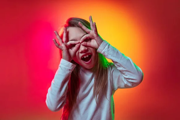 Jedna legrační školačka, šťastný kluk dělat obličeje izolované na červeném žlutém pozadí studia v neonovém světle, filtr. — Stock fotografie