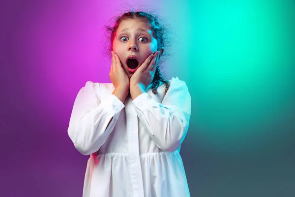Studio záběr šokované, vyděšené holčička, dítě stojící s otevřenými ústy izolované na gradient fialové modré barvy pozadí v neonovém světle — Stock fotografie