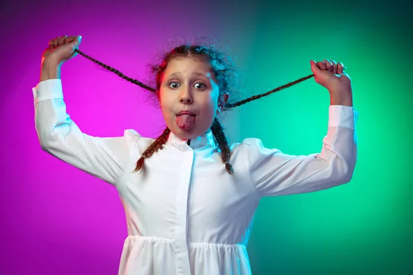 Retrato de meio comprimento de menina engraçada, criança fazendo rostos isolados no gradiente roxo azul cor de fundo em luz de néon — Fotografia de Stock