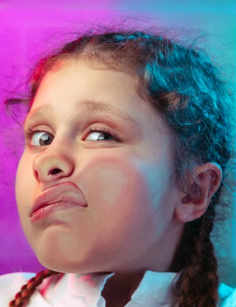 Primer plano niña, lindo niño apoyado contra el vidrio transparente por la mejilla aislado sobre fondo de estudio púrpura azul en luz de neón — Foto de Stock