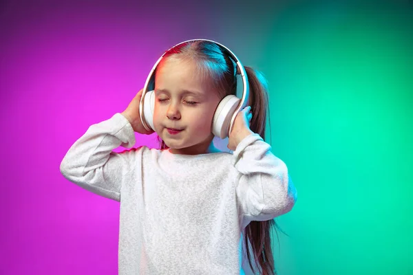 Половина длиной портрет спокойной маленькой девочки, ребенок слушает аудио сказочный хвост в наушниках изолированы на градиент фиолетовый синий цвет фона в неоновом свете — стоковое фото