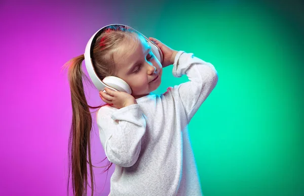 One beautiful little girl, kid listening music in headphones isolated on gradient purple blue color background in neon light — Stock Photo, Image