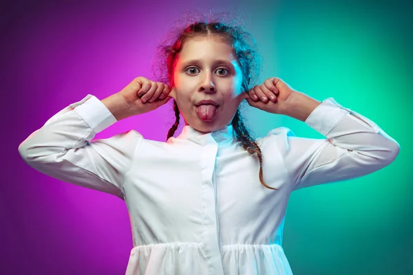 Half-length portrait of funny little girl, kid making faces isolated on gradient purple blue color background in neon light Royalty Free Stock Images