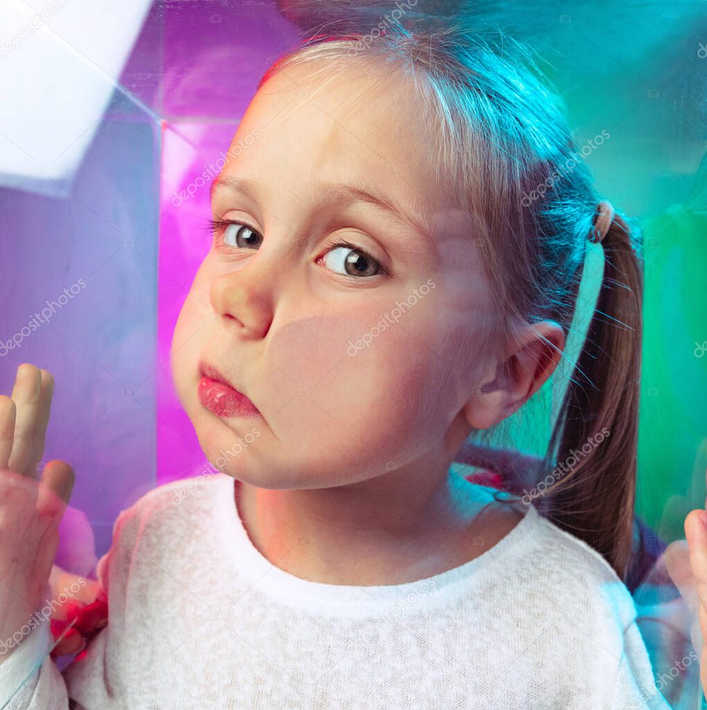 Close-up little girl, cute kid leaning against transparent glass by cheek isolated on blue purple studio background in neon light