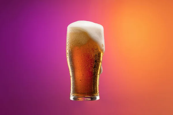 Plná sklenice zpěněného lehkého ležáckého piva izolovaného nad svahem fialové a oranžové barvy pozadí v neonu. Koncept alkoholu, nápojů a festivalů. — Stock fotografie