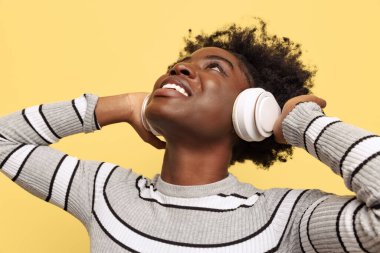 Sarı arka planda izole edilmiş beyaz kulaklıklı, koyu tenli, çekici bir kız. Duyguların kavramı