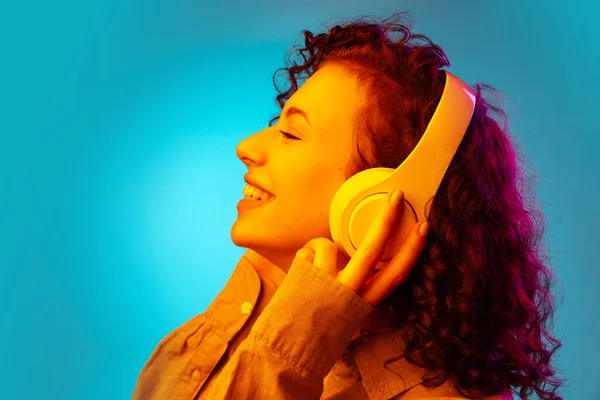 Widok z boku podekscytowany młoda piękna dziewczyna w słuchawki słuchanie muzyki, książki audio izolowane na niebieskim tle w neonie — Zdjęcie stockowe