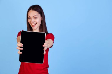 Mavi stüdyo arka planında izole edilmiş dijital tablet kullanan kırmızı tişörtlü duygusal genç bir kız. Duyguların kavramı