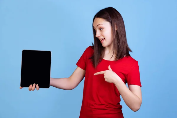 Emocionální mladá krásná dívka v červeném tričku pomocí digitálního tabletu izolované na modrém pozadí studia. Pojetí emocí — Stock fotografie