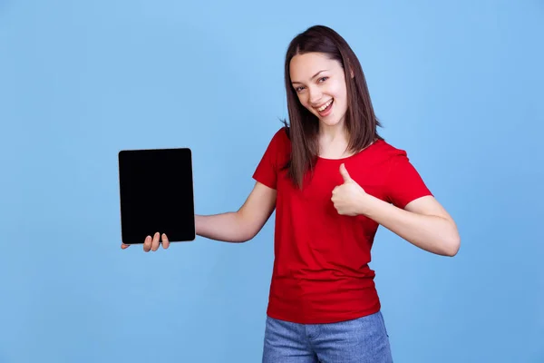 Emocionální mladá krásná dívka v červeném tričku pomocí digitálního tabletu izolované na modrém pozadí studia. Pojetí emocí — Stock fotografie