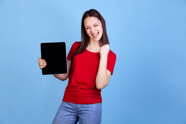 Emocjonalna młoda piękna dziewczyna w czerwonym t-shircie z cyfrowym tabletem odizolowanym na niebieskim tle studia. Pojęcie emocji — Zdjęcie stockowe