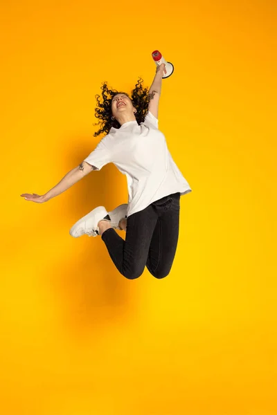 Full längd porträtt av ung glad flicka, student i vit t-shirt och jeans hoppa isolerad på gul bakgrund. — Stockfoto