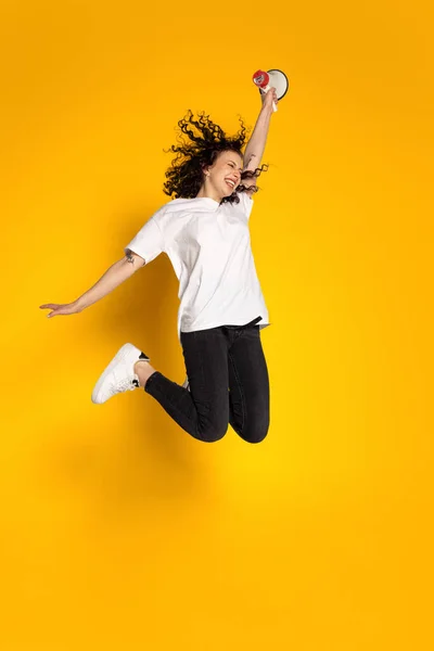 Full längd porträtt av ung glad flicka, student i vit t-shirt och jeans hoppa isolerad på gul bakgrund. — Stockfoto