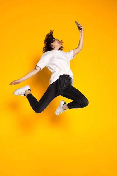Retrato completo de jovem menina alegre, estudante de t-shirt branca e jeans pulando isolado no fundo amarelo. — Fotografia de Stock