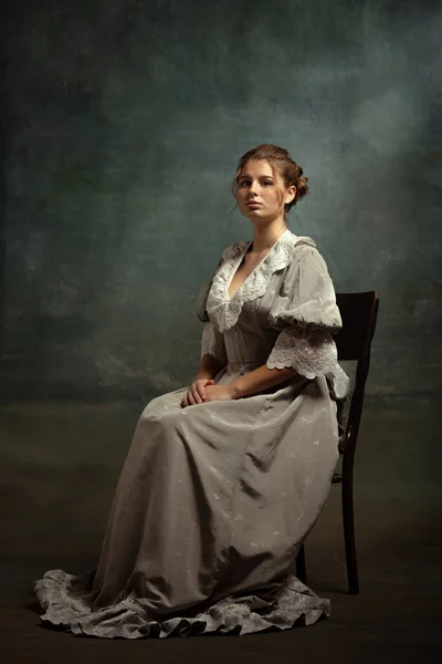 Traurige junge schöne Mädchen in grauem Kleid mittelalterlichen Stils sitzt auf Stuhl isoliert auf dunklem Vintage-Hintergrund. Epochenvergleich Konzept, flämischer Stil. — Stockfoto