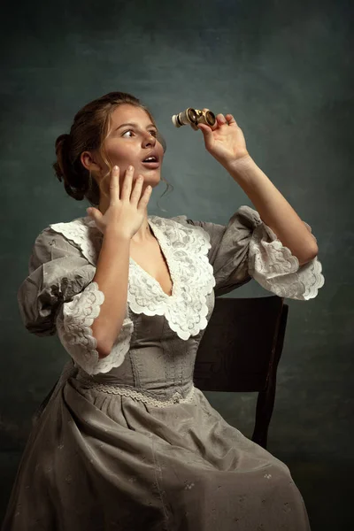 Surprised young beautiful girl in gray dress of medieval style using opera glasses isolated on dark background. — Stock Photo, Image