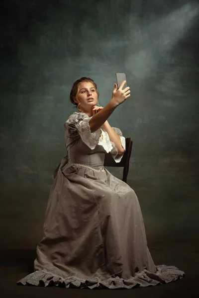 Retrô retrato de jovem menina romântica em vestido cinza de estilo medieval usando telefone isolado no fundo escuro do vintage. — Fotografia de Stock
