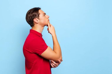 Profil görünümlü yakışıklı bir genç, geleceği düşünen bir öğrenci mavi stüdyo arka planında izole edilmiş.